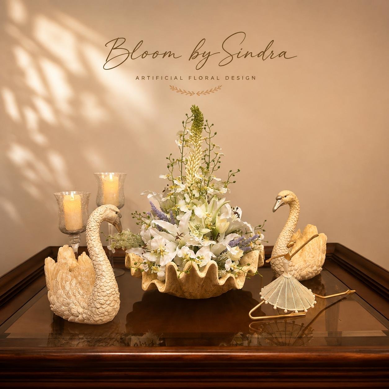 Bloom by Sindra branded photo of white lily and orchid arrangement in a clamshell dish flanked by two swan figurines
