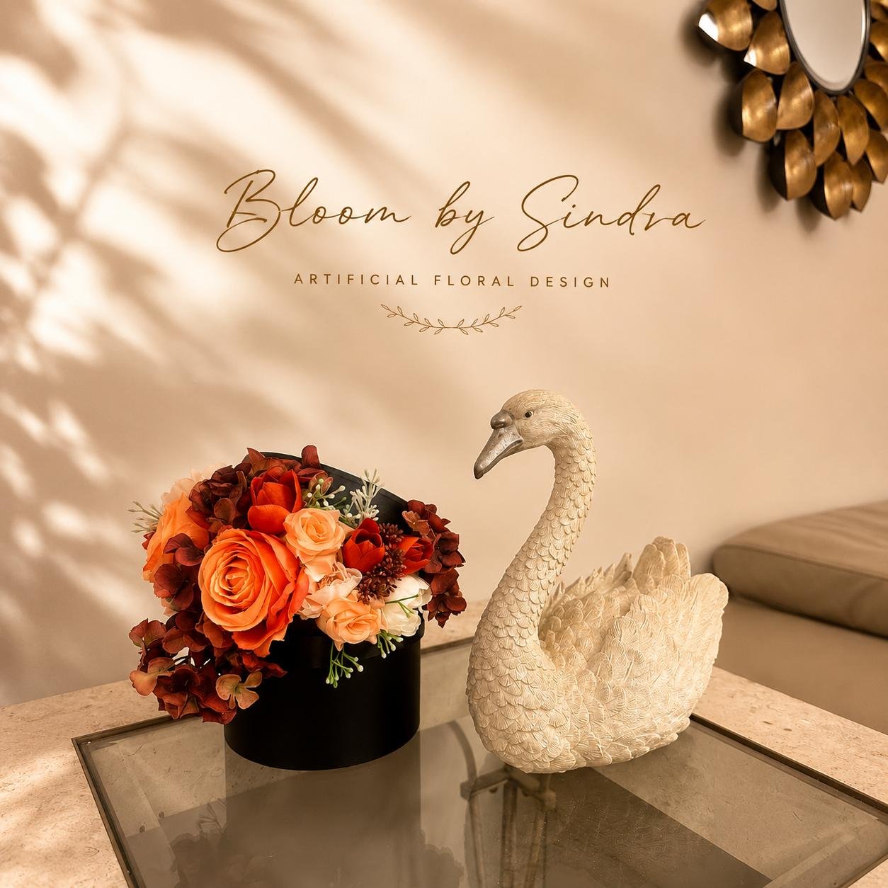 Bloom by Sindra branded photo of a black box vase filled with orange roses and dark florals beside a swan figurine